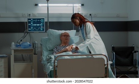 Doctor Installing Oximeter On Patient Hand In Hospital Ward At Medical Clinic. African American Medic Doing Consultation On Sick Old Man With Nasal Oxygen Tube And IV Drip Bag For Disease