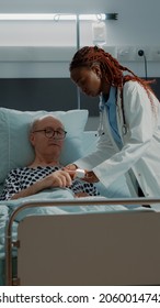 Doctor Installing Oximeter On Patient Hand In Hospital Ward At Medical Clinic. African American Medic Doing Consultation On Sick Old Man With Nasal Oxygen Tube And IV Drip Bag For Disease