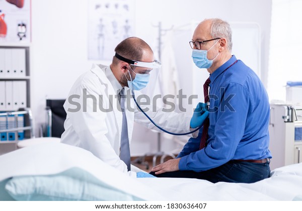 Doctor listening heart beat\
of senior man with stethoscope during examination on hospital room\
to give a diagnosis and using protection mask for\
covid-19.