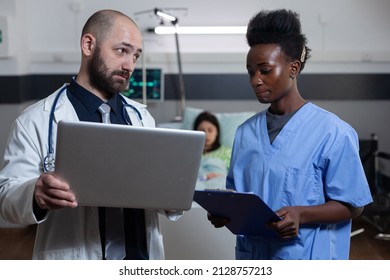 Doctor Looking Judgemental At Hospital Nurse Feeling Guilty For Getting Lab Results Wrong Standing In Patient Ward. Medic Holding Laptop Computer Showing Diagnostic To Health Care Worker.