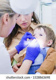 The Doctor Looks At The Teeth Of A Small Child. The Baby Is Sitting In The Arms Of Mom. Dentist Examining Boy's Teeth In Clinic. A Small Patient In The Dental Chair Smiles. Dantist Treats Teeth. Close