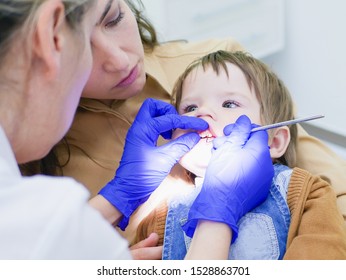 The Doctor Looks At The Teeth Of A Small Child. The Baby Is Sitting In The Arms Of Mom. Dentist Examining Boy's Teeth In Clinic. A Small Patient In The Dental Chair Smiles. Dantist Treats Teeth. Close