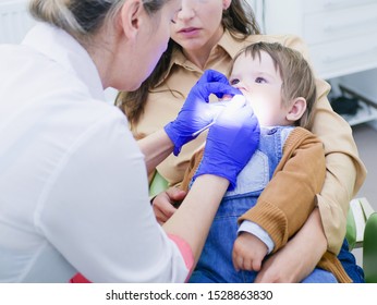 The Doctor Looks At The Teeth Of A Small Child. The Baby Is Sitting In The Arms Of Mom. Dentist Examining Boy's Teeth In Clinic. A Small Patient In The Dental Chair Smiles. Dantist Treats Teeth. Close