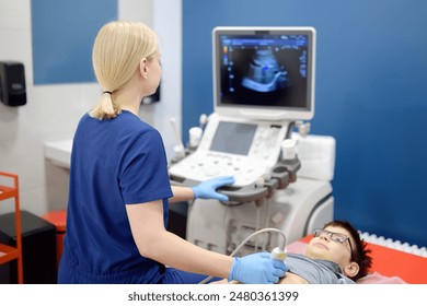 Doctor making abdominal ultrasound for boy using scanner machine. Female doc runs ultrasound sensor over patient tummy looking at screen. Medical examination of internal organs for school kids - Powered by Shutterstock