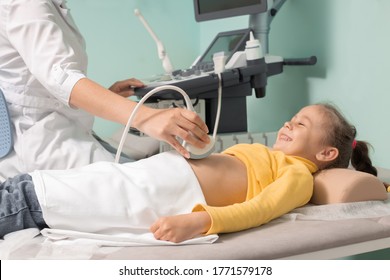 Doctor Making Abdominal Ultrasound For Child Girl Using Scanner Machine, Side View. Woman Runs Ultrasound Sensor Over Patient Tummy Looking At Screen. Diagnostic Examination Of Internal Organs.