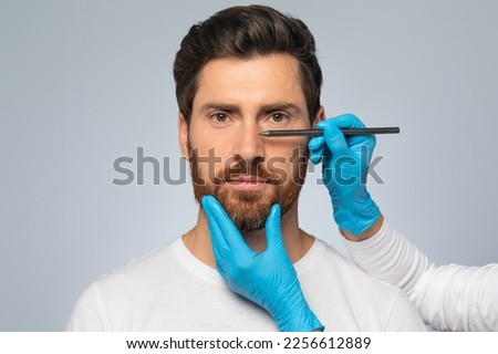 Doctor making marks on male patient's face, middle aged man on consultation at surgeon, standing on grey background. Facial plastic surgery concept