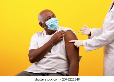 Doctor Making Vaccine Shot Against Coronavirus To Elderly Black Male Patient On Orange Studio Background. African American Man Wearing Medical Mask, Getting Vaccinated From Covid