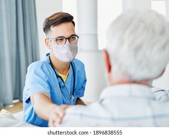 Doctor Or Male Caregiver With Senior Man Wearing Protective Mask At Home Or Nursing Home