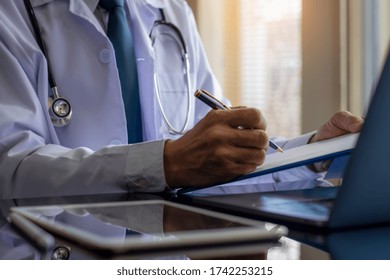 Doctor Man In White Lab Coat Writing Note On Data Chart Clipboard With Laptop Computer And Digital Tablet On The Desk At Clinic Or Hospital. Online Medical Learning Or Working Concept.