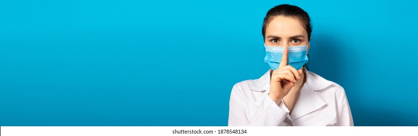 The doctor in the mask shows a sign of silence finger at the mouth. Medical mystery in medicine and health care, medical error and secrets of new medical research. Medical secrecy. Banner blue copy
