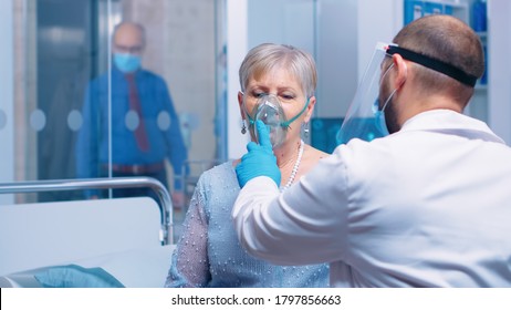 Doctor in mask and visor helping old lady to breathe with a respiratory oxygen mask while sitting on hospital bed in modern private clinic. Coronavirus covid-19 outbreak healthcare crisis.