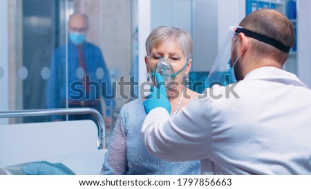 Doctor in mask and visor helping old lady to breathe with a respiratory oxygen mask while sitting on hospital bed in modern private clinic. Coronavirus covid-19 outbreak healthcare crisis.