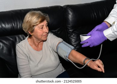 A Doctor Measures A Senior Woman's Blood Pressure With A Blood Pressure Cuff On Her Arm While She Is Sitting On A Couch. Concept Of Home Health Care