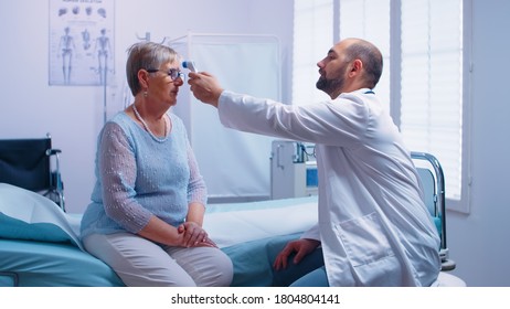 Doctor measuring temperature to senior woman who sits on hospital bed. Using digital infrared contactless thermometer in modern private clinic. Medical examination for infections and disease