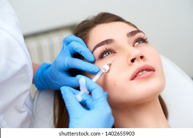 Doctor In Medical Gloves With Syringe Injects Botulinum Under Eyes For Rejuvenating Wrinkle Treatment. Filler Injection For Eye Wrinkles Smoothing. Plastic Aesthetic Facial Surgery In Beauty Clinic