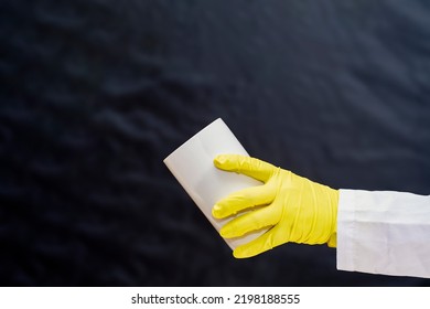 A Doctor In A Medical Gown And Protective Gloves Holds In His Hand A Large White Container Without Inscriptions. The Face Is Not Visible. Empty Template For Text.