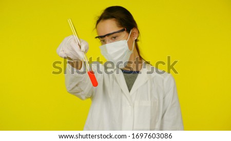 Doctor in a medical mask, goggles and latex gloves looks at the tests in the flask. A young girl in a white coat on a yellow background holds hand a flask with red liquid
