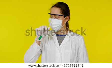 Doctor in a medical mask, goggles and latex gloves looks at the green liquid in the test tube. Young girl in a white coat on a yellow background
