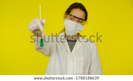 Doctor in medical mask, goggles, latex gloves looks at the tests in the tube. Young girl in white coat on yellow background holds hand tube with green liquid