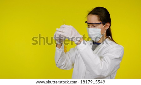 Doctor in medical mask goggles latex gloves looks at the tests in the tube. Woman in white coat on yellow background holds hands tube with gold yellow liquid