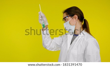 Doctor in medical mask goggles latex gloves looks tests in tube. Woman in white coat on yellow background holds hand tube with green liquid