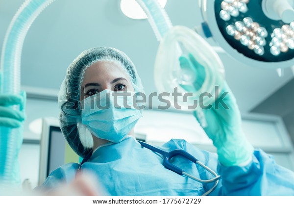 Doctor in medical mask and protective\
clothes standing with an anesthesia mask in her\
hand