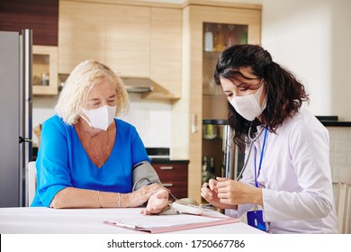 Doctor In Medical Mask Visiting Female Senior Patient, Checking Her Blood Pressure And Filling Medical History