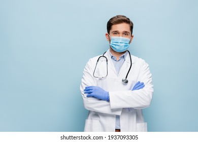 doctor in medical mask and white coat with stethoscope standing with crossed arms isolated on blue