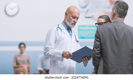 Doctor Meeting A Couple In The Hospital Aisle, He Is Checking Medical Records, Medicine And Healthcare Concept