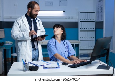 Doctor And Nurse Doing Teamwork For Medical Checkup At Office. Medical Team Using Computer And Tablet For Healthcare System, Working Late At Night. Woman Assistant Looking At Medic.