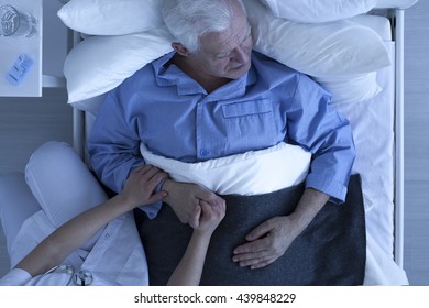 Doctor Or Nurse Holding Hand Of Senior Male Patient Lying In Hospice Bed