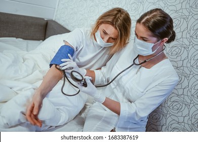 Doctor Nurse In Protective Face Mask Measures Blood Pressure With A Sphygmomanometer. Coronavirus (COVID-19) Concept. First Symptoms. Woman Sick Of Flu Viral Infection In Home Isolation Quarantine