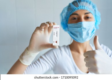 Doctor Or Nurse In Protective Face Mask And Gloves Doing Positive Gesture With Hand, Thumbs Up, Holding Negative COVID-19 Test. Diagnosis Of Coronavirus, Inhibition Of Disease Outbreaks.