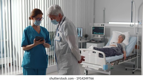 Doctor And Nurse In Safety Mask Discussing Diagnosis On Digital Tablet With Woman Patient Lying In Bed On Background. Oncologist And Nurse Watching Mri Scan On Tablet Of Woman With Cancer