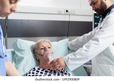 Doctor And Nurse Supervising Senior Woman Breathing With Oxygen Mask, Laying In Bed Because Of Pulmonary Disease. Patient Laying In Bed With Iv Dip Bag Attached.