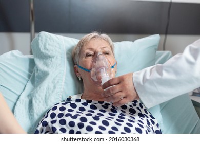 Doctor And Nurse Supervising Senior Woman Breathing With Oxygen Mask, Laying In Bed Because Of Pulmonary Disease. Patient Laying In Bed With Iv Dip Bag Attached.