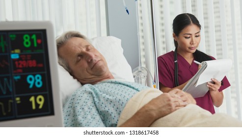 Doctor or nurse taking notes on chart with senior Caucasian male patient recovering in hospital bed in foreground. Japanese woman medical professional monitoring recovery - Powered by Shutterstock