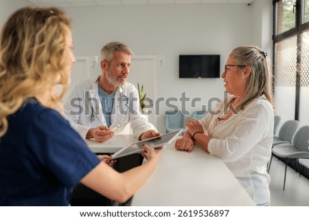 Doctor and nurse using tablet while discussing with a senior patient in the waiting room of a modern clinic, providing healthcare assistance and showing medical information