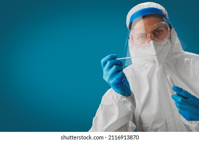 A Doctor Or Nurse Wearing A Hazmat Suit Or PPE With A Face Shield, White N95 Respirator And Blue Gloves Takes A Sample Of A COVID-19 PCR Test Using A Cotton Swab On A Blue Background. Copy Space.