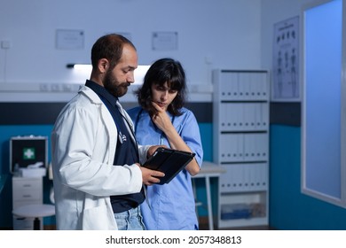 Doctor And Nurse Working Late At Night For Healthcare Using Digital Tablet. Medical Assistant And Medic Doing Teamwork At Office For Medicine. Woman And Man Looking At Modern Gadget