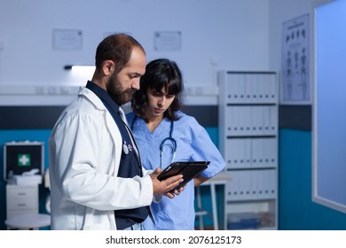 Doctor And Nurse Working Late At Night For Healthcare Using Digital Tablet. Medical Assistant And Medic Doing Teamwork In Cabinet For Medicine. Specialists Looking At Modern Device