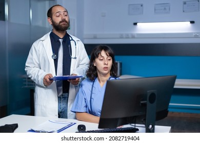 Doctor And Nurse Working With Technology On Computer For Medical Care And Treatment. Team Of Workers Using Monitor And Checkup Files For Healthcare System, Working Late At Night.