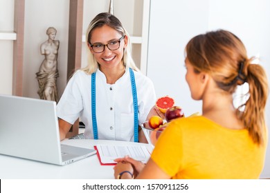 Doctor Nutritionist, Dietician And Female Patient On Consultation In The Office. Young Smiling Female Nutritionist In The Consultation Room. Nutritionist Desk With Healthy Fruit And Measuring Tape.