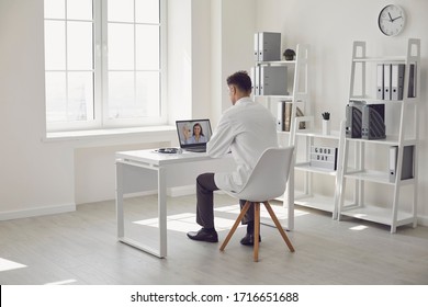 Doctor Online. Male Doctor Works Talking Video Call With Patient Using Bast Shoe While Sitting At Desk In Back View Clinic Office.