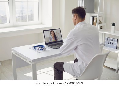 Doctor Online. Male Doctor Works Talking Video Call With Patient Using Bast Shoe While Sitting At Desk In Back View Clinic Office.