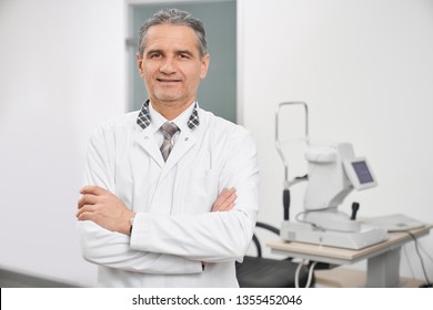 Doctor Ophthalmologist Wearing In White Medical Coat Standing In Medical Room Of Clinic. Eye Specialist Posing With Hands Crossed And Looking At Camera. Slit Lamp, Special Equipment On Background.