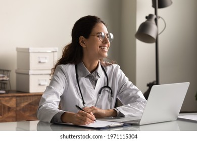Doctor at paperwork. Happy young latin female medical worker in glasses sit at workplace in clinic office look aside dreaming. Smiling millennial woman gp distracted from taking notes records thinking