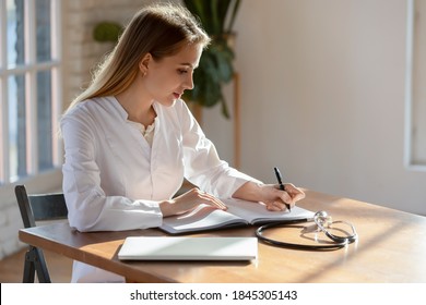 Doctor At Paperwork. Responsible Concentrated Millennial Woman Family Therapist Sitting At Desk In Hospital Cabinet Making Recordings To Paper Casebook, Writing Medical History Or Treatment Profile