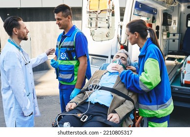 Doctor And Paramedics Checking Health Of Mature Male With Breathing Mask Lying In Ambulance Gurney Brought To Hospital By EMT Workers. Emergency Medical Services