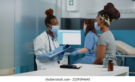 Doctor And Patient With Face Masks Talking About Disease At Annual Checkup Visit For Examination. Medic Consulting Young Woman In Cabinet While Having Protection Against Coronavirus